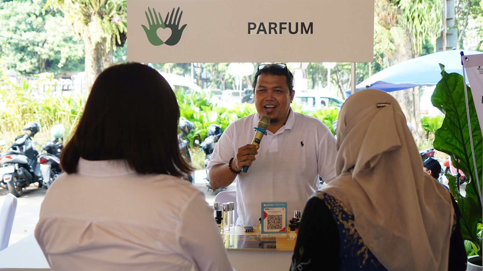 Alumni Wirausaha Menjelaskan Produk Parfum Racikan Mandiri Pelaku usaha alumni binaan parfum YIIM memegang mikrofon mempromosikan produk parfum di meja pameran