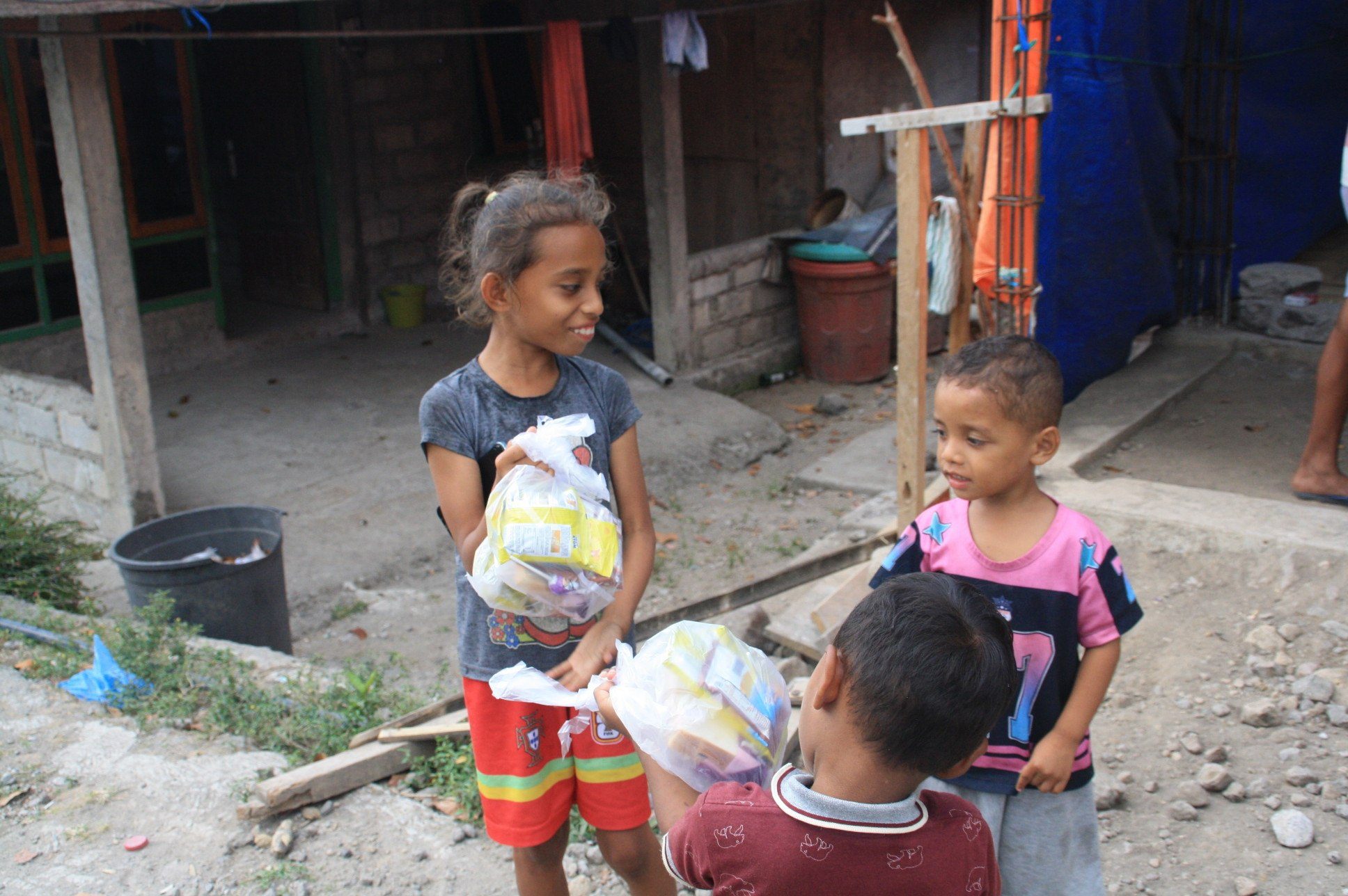 Keceriaan Anak-Anak Menerima Bantuan Pangan Darurat Pasca Gempa NTT Tiga anak tersenyum ceria memegang bungkusan bantuan makanan ringan dan nutrisi di halaman rumah