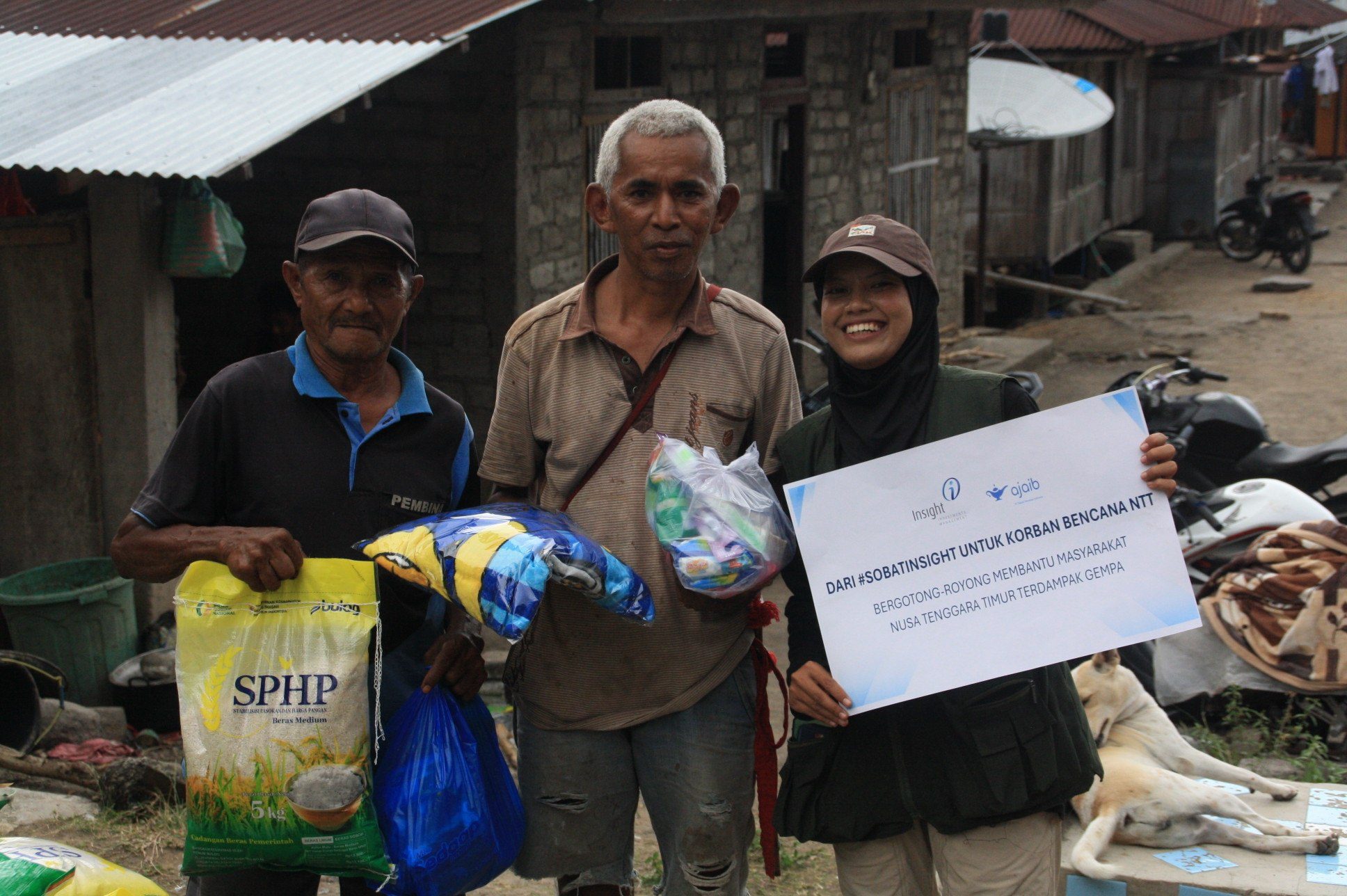 Penyerahan Bantuan Beras dan Family Kit Dukungan Sobat Insight untuk Korban Gempa NTT Dua warga penerima manfaat memegang bantuan beras Bulog, selimut, dan hygiene kit bersama relawan pembawa plakat